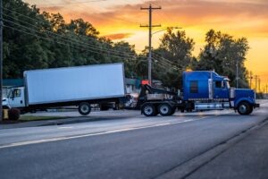 A blue tow truck hauls a damaged semi-truck with a trailer after an accident on a local road.