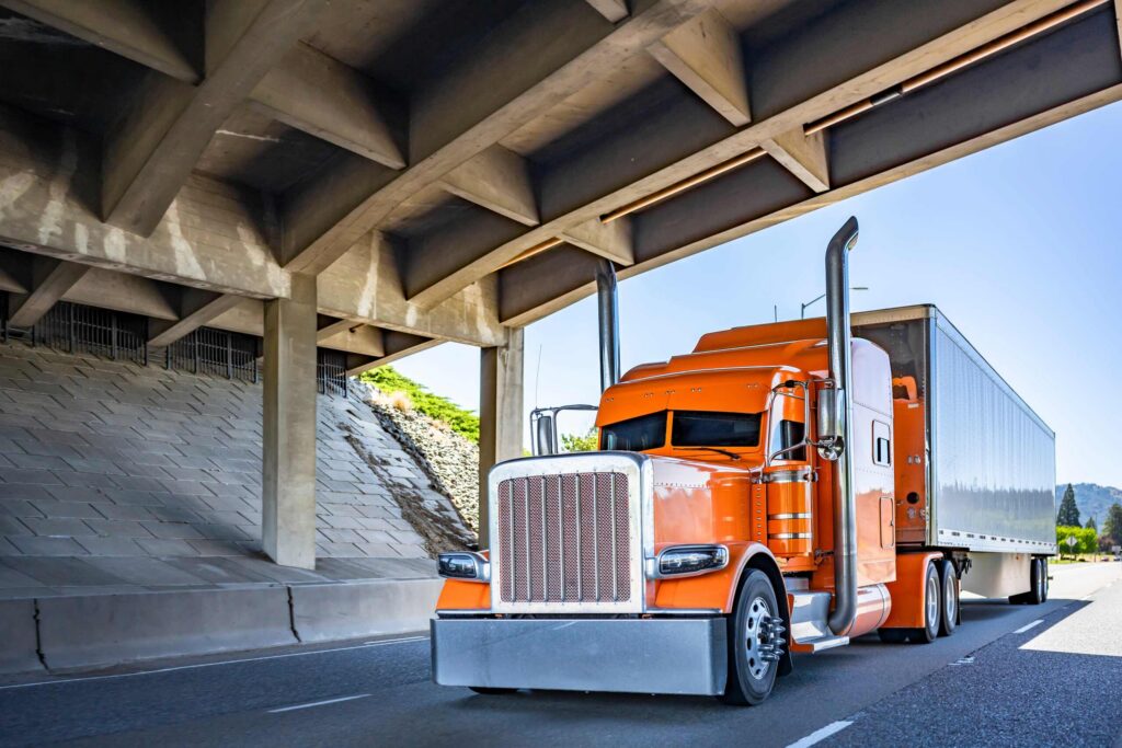 An orange semi-truck driving on a highway under a concrete bridge overpass.
