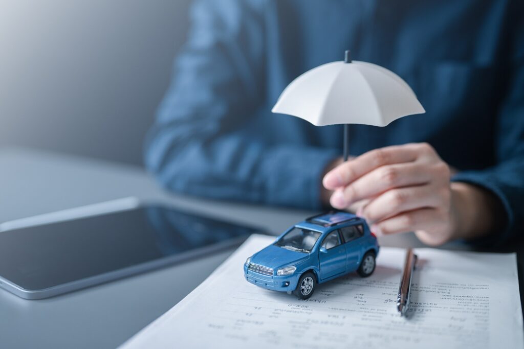 A person holding a small umbrella over a toy blue car on insurance documents, representing coverage claims managed by Boca Raton Ride Share Accident Lawyers.
